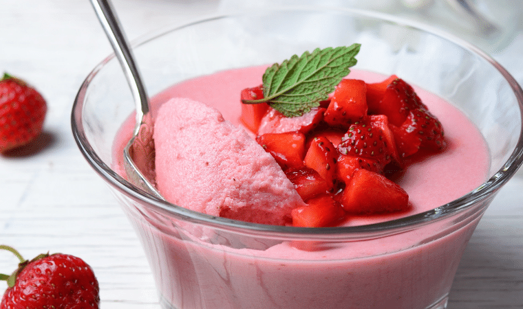 Receita de Mousse de Morango servida em uma taça individual de vidro com pedaços de morangos por cima com uma colher tirando uma porção da mousse