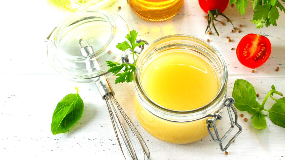 molho para salada com mostarda pronto em uma jarra de vidro com um garfo de salada ao lado e com folhas verdes decorando a mesa e um pequeno tomate cereja partido ao meio